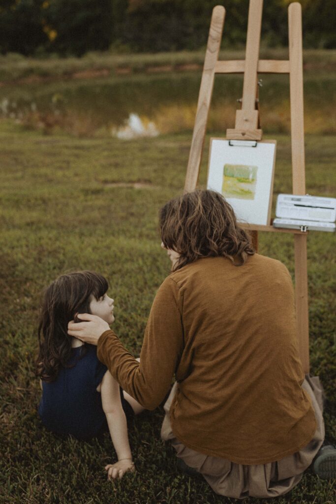 Mother embraces daughter while painting a pond landscape outdoors, capturing a quiet, emotive moment of connection and creativity.
