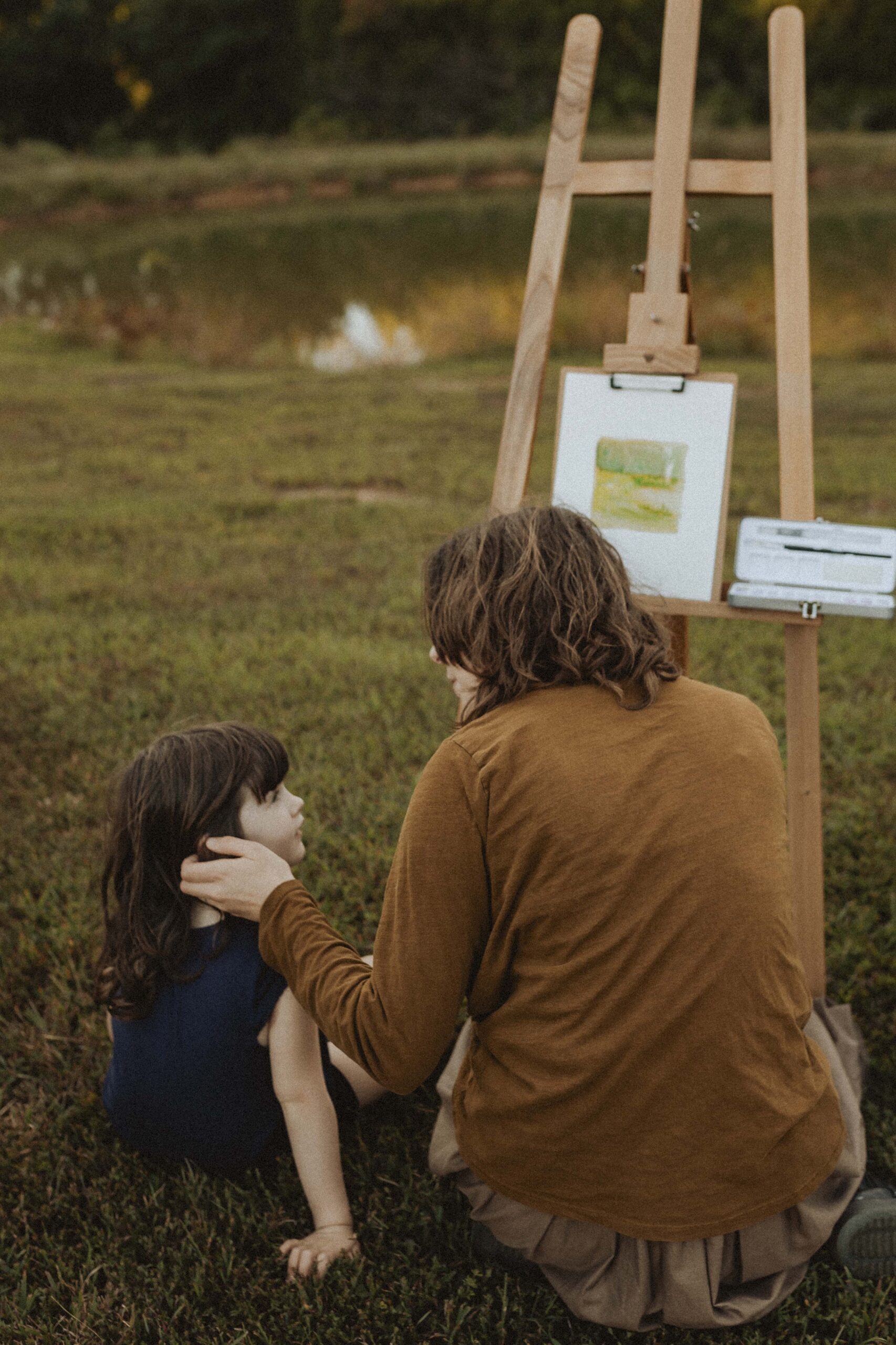 Mother embraces daughter while painting a pond landscape outdoors, capturing a quiet, emotive moment of connection and creativity.