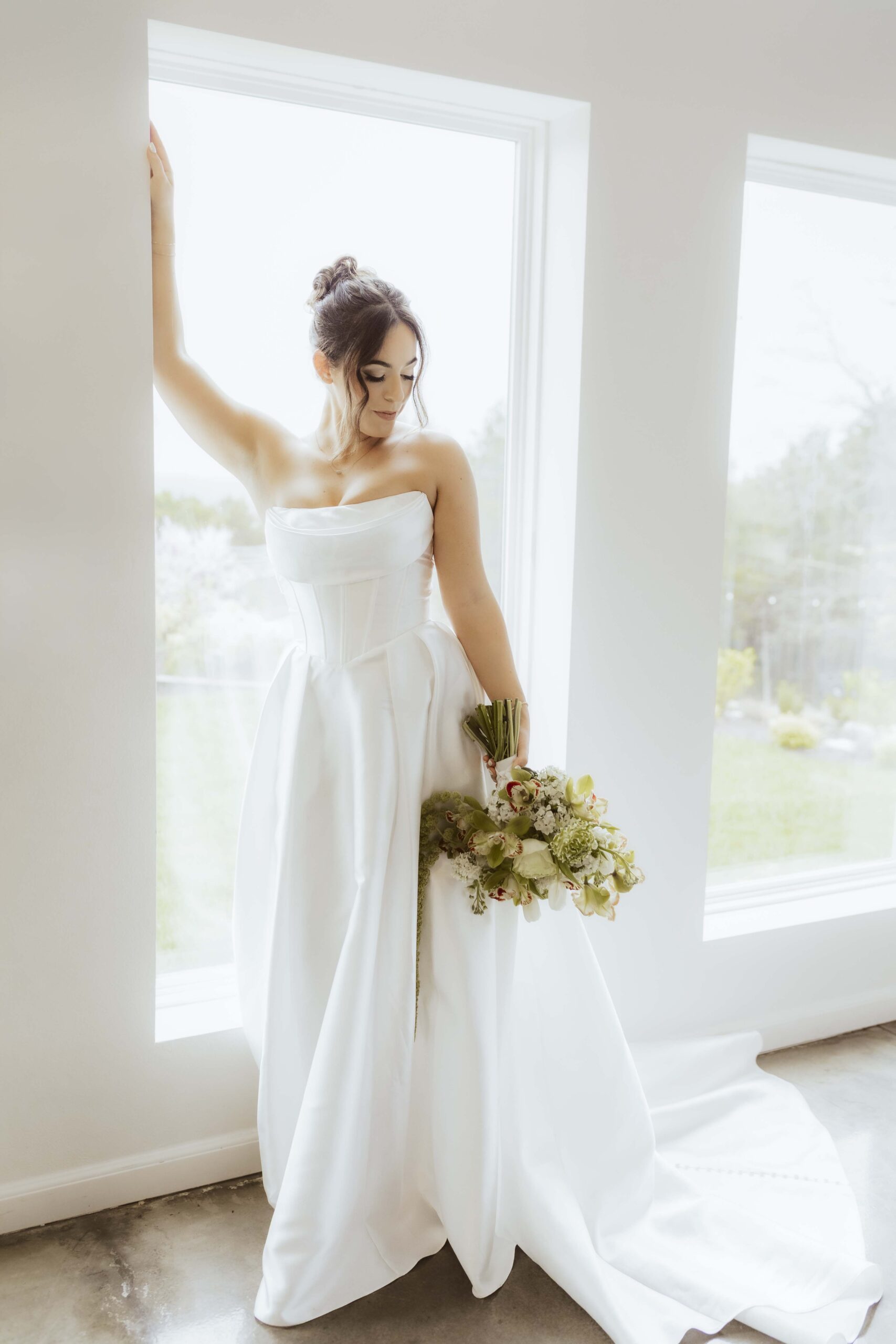 Bride stands in front of large modern windows, bathed in natural light, holding a lush green bouquet in an editorial-inspired pose.