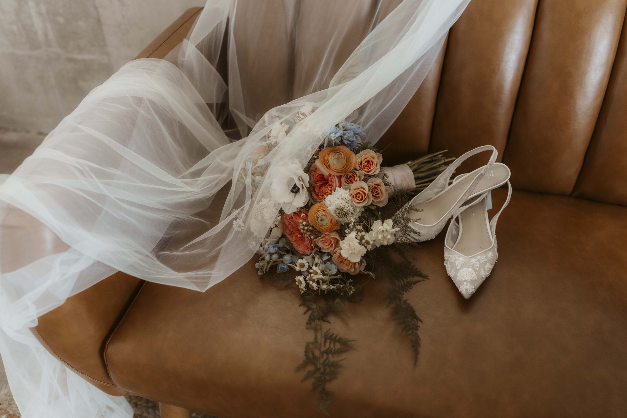 Flat lay of a bride’s veil, bouquet, and heels arranged on a rich leather couch in soft, natural light—capturing timeless elegance.