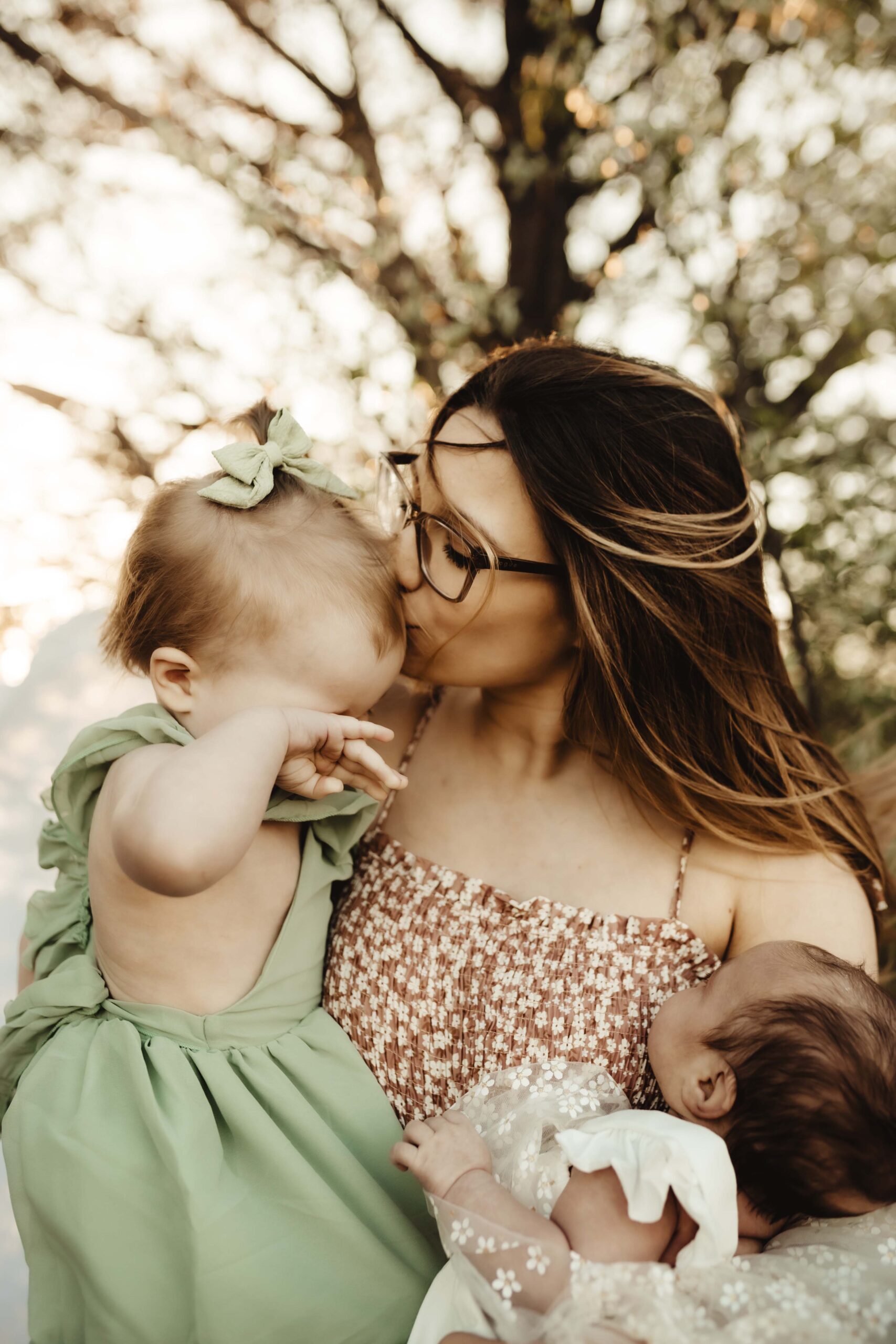 Mother holds her toddler and infant during a family session; wind lifts her hair as she kisses her tired toddler, who rubs her eyes in a quiet, emotional moment.