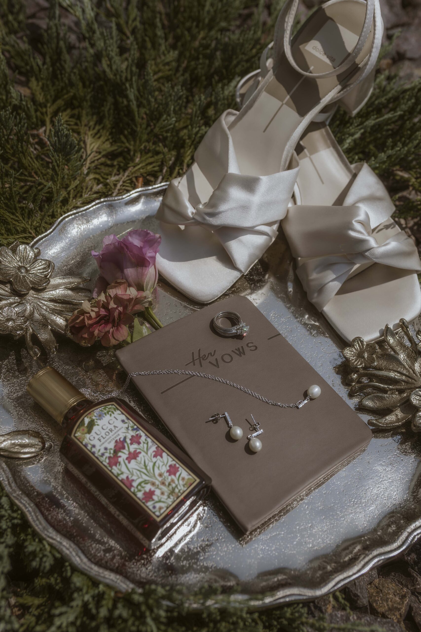 Bridal detail photo with ivory heels, Gucci Flora perfume, pearl jewelry, her vow book, and wedding rings on a vintage silver tray.